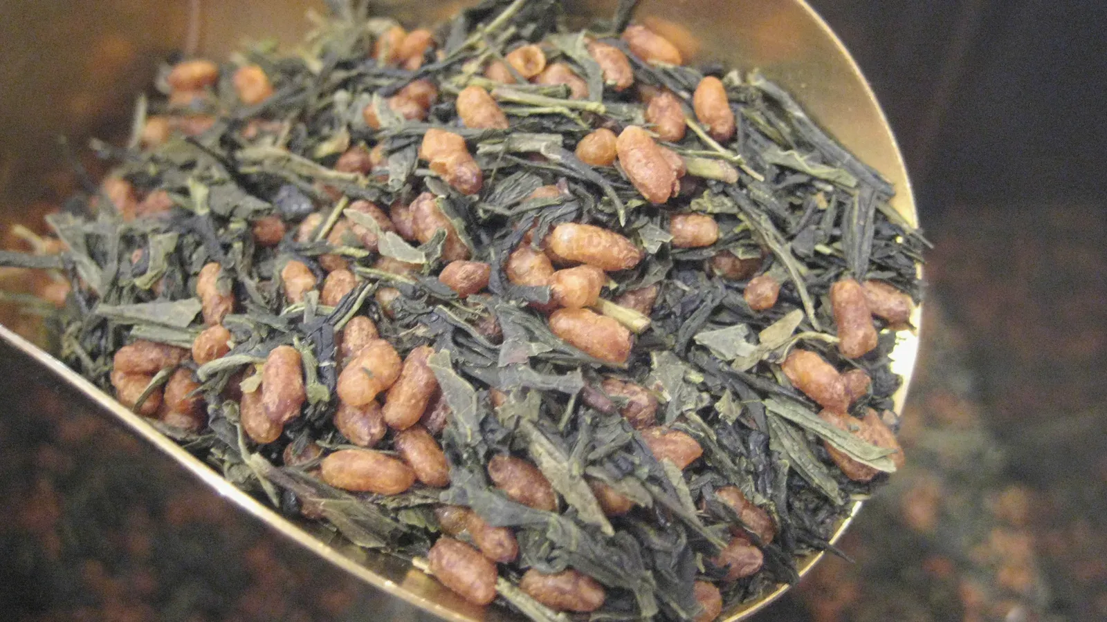 A brass scoop of genmaicha — green tea needles mixed through pale toasted rice grains